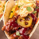 Close-up of a slow cooker BBQ beef taco with coleslaw, jalapeños, and BBQ sauce on a soft flour tortilla.