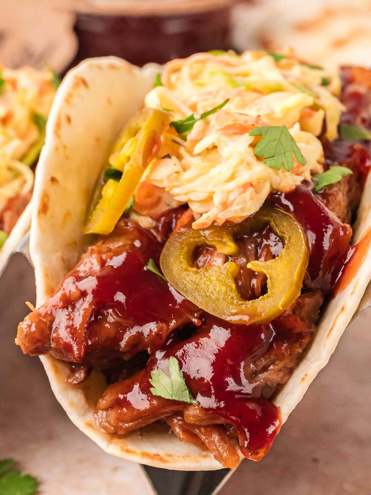 Close-up of a slow cooker BBQ beef taco with coleslaw, jalapeños, and BBQ sauce on a soft flour tortilla.