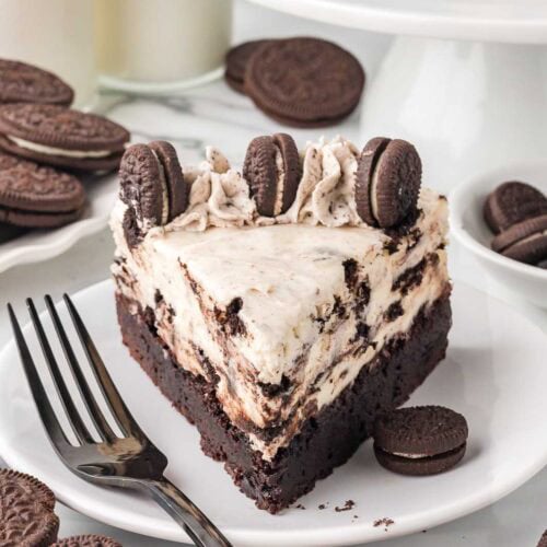 Oreo cheesecake slice on a plate with brownie base, Oreo whipped topping, and cookies on top.
