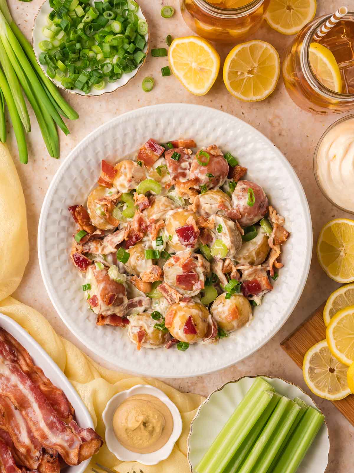 Creamy potato salad with bacon, celery, and green onions served in a bowl with fresh ingredients around it.