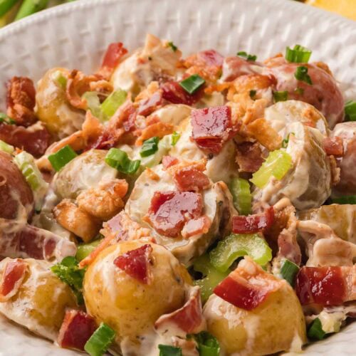 Potato salad with bacon and green onions served in a white bowl with fresh ingredients in the background.