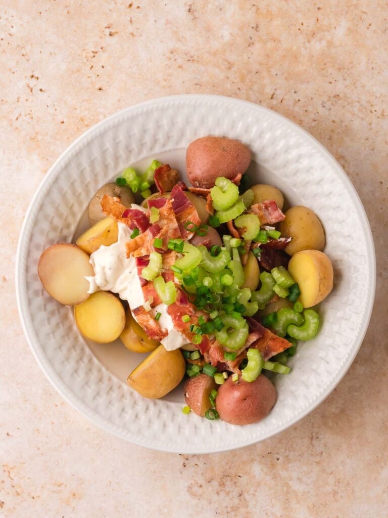 Baby potatoes topped with bacon, celery, green onions, and dressing before mixing.

