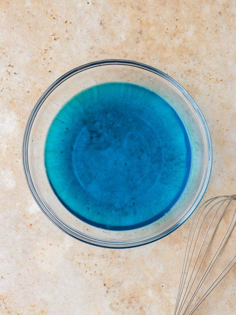 Blue Jello mixture in a glass bowl on a countertop with a whisk beside it.