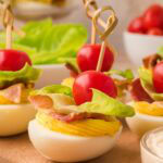 BLT deviled eggs with creamy filling, bacon, lettuce, and tomato on a wooden serving board.
