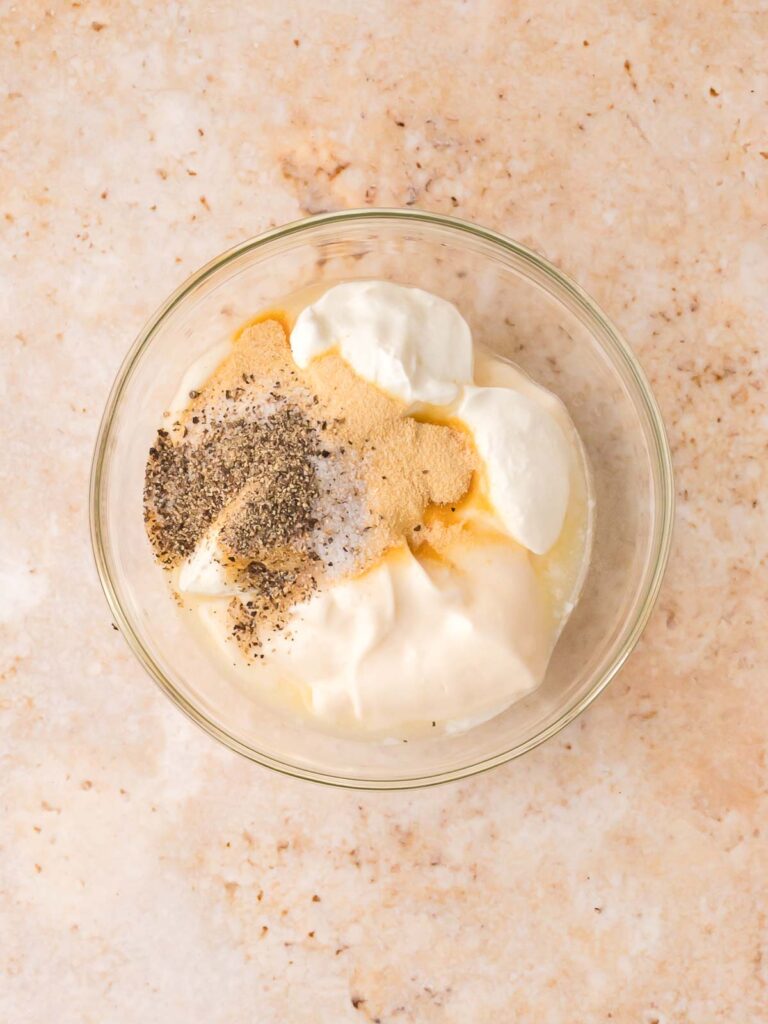 Mayonnaise, sour cream, lemon juice, and seasonings in a glass bowl for pasta salad dressing.