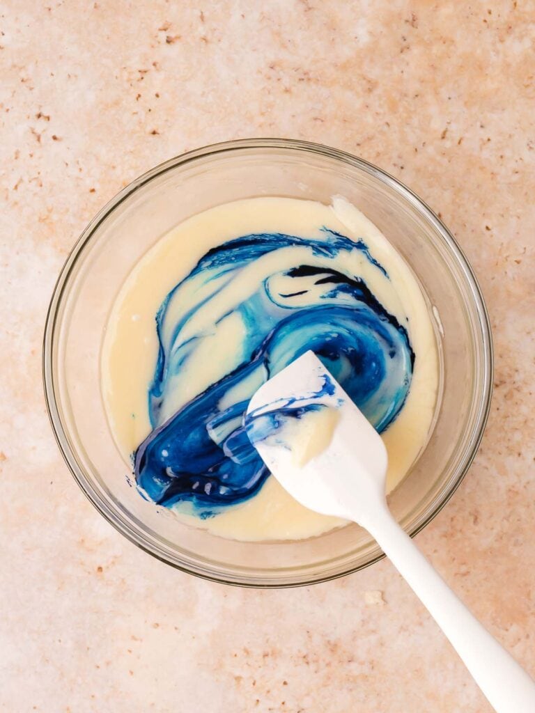Blue food coloring being stirred into melted white chocolate fudge mixture.