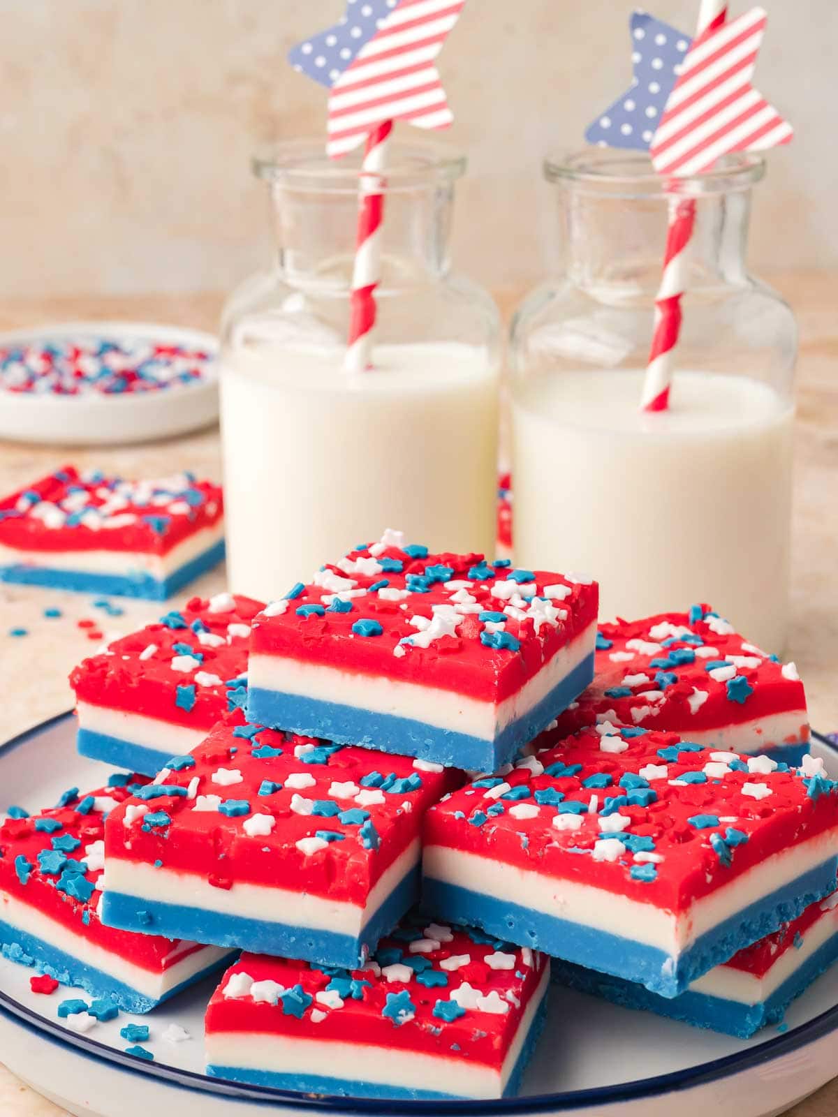 Red white and blue layered fudge squares stacked on a plate with patriotic sprinkles and milk bottles in the background.