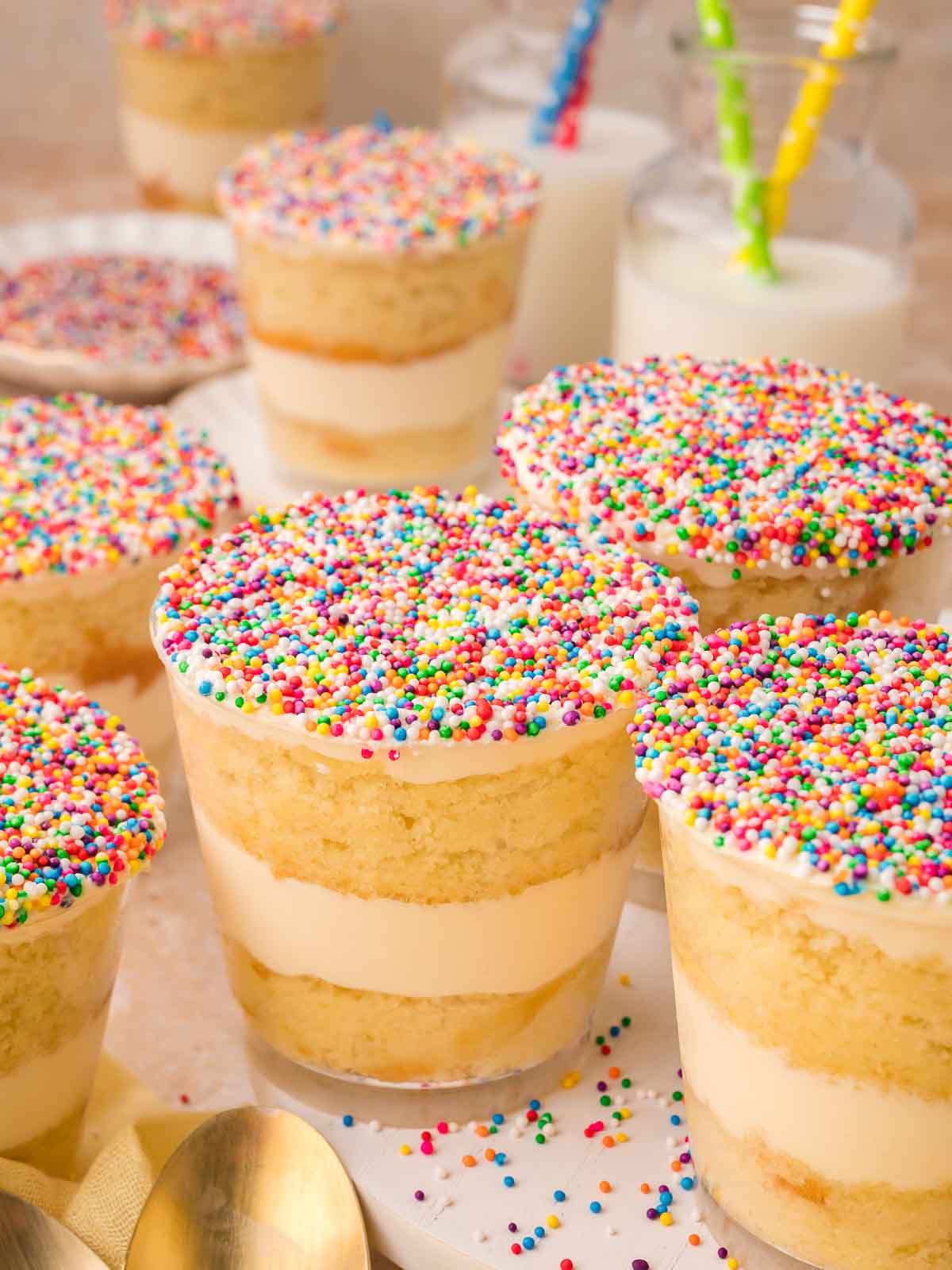 Several finished dot cakes with rainbow nonpareil tops arranged on a white surface with gold spoons and glasses of milk by Tessie's Table.