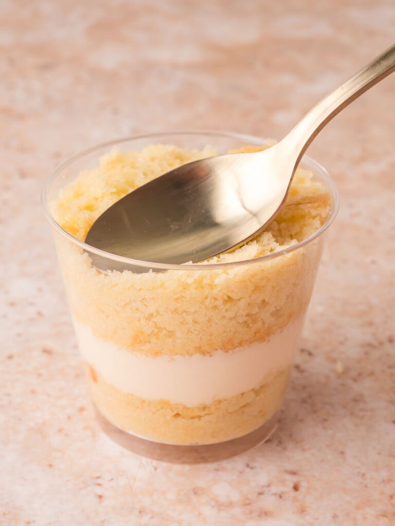 Layered vanilla cake and buttercream in a clear plastic cup being smoothed with a gold spoon for dot cakes by Tessie's Table.