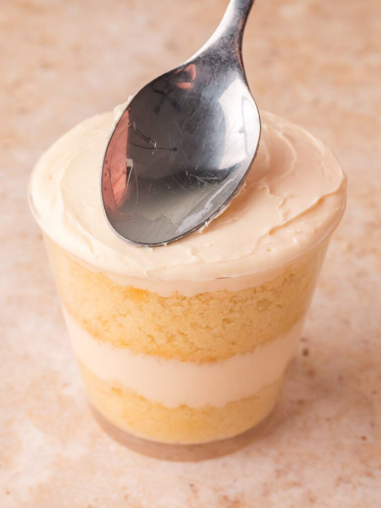 Buttercream frosting being smoothed flat and flush with the rim of a clear cup of layered dot cakes by Tessie's Table.
