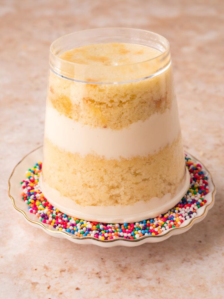 Assembled dot cake in a clear cup showing vanilla cake and buttercream layers, sitting on a plate of rainbow nonpareils by Tessie's Table.