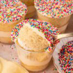 Spoonful of vanilla cake and buttercream lifted from a dot cake cup surrounded by more finished dot cakes topped with rainbow nonpareils by Tessie's Table.