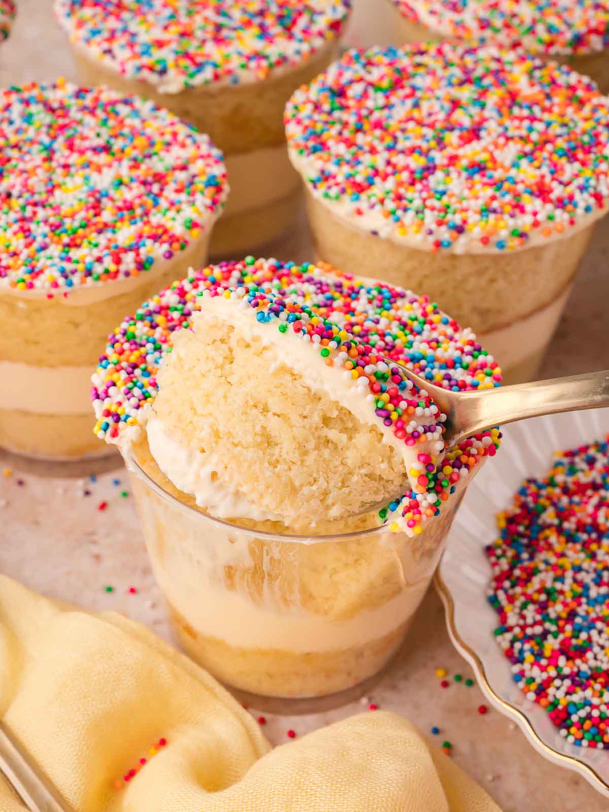 Spoonful of vanilla cake and buttercream lifted from a dot cake cup surrounded by more finished dot cakes topped with rainbow nonpareils by Tessie's Table.