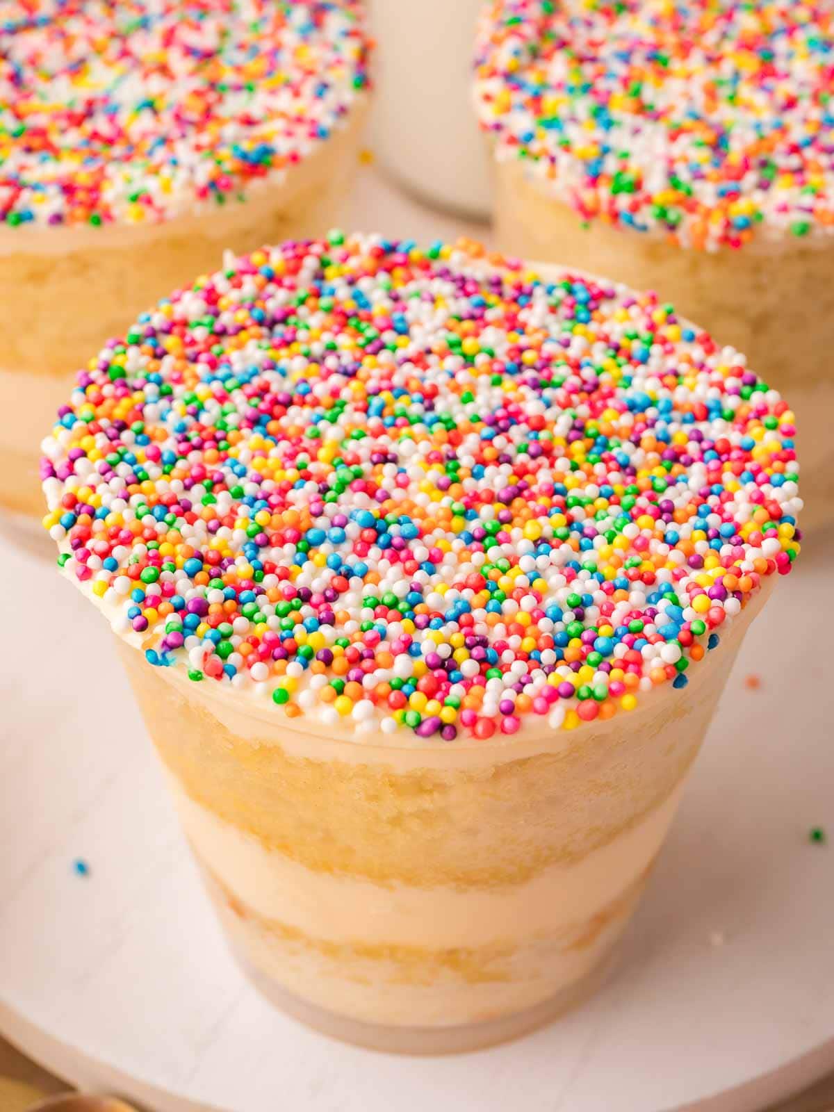 Close-up of a finished dot cake in a clear cup topped with thick rainbow nonpareils, showing vanilla cake and buttercream layers, by Tessie's Table.