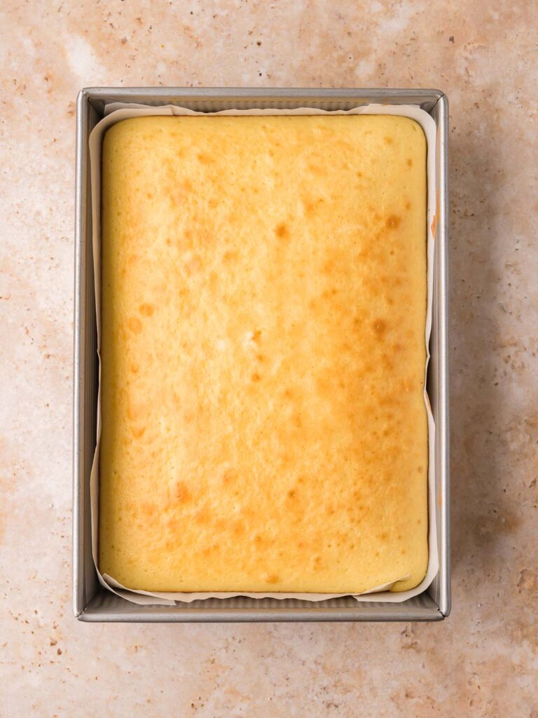 Baked vanilla cake in a parchment-lined 9x13 pan, golden on top, cooling for dot cakes by Tessie's Table.