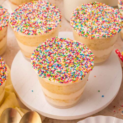 Multiple dot cakes topped with rainbow nonpareils displayed on a white cake stand with glasses of milk in the background by Tessie's Table.