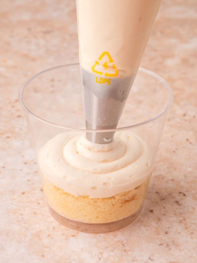 Piping buttercream frosting over the first cake round in a clear plastic cup for dot cakes by Tessie's Table.
