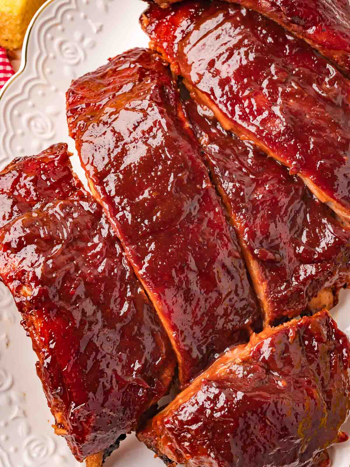 Close-up of sliced fall-off-the-bone oven BBQ ribs with a deep red sticky glaze on a white decorative plate — Fall-Off-the-Bone Oven BBQ Ribs by Tessie's Table.