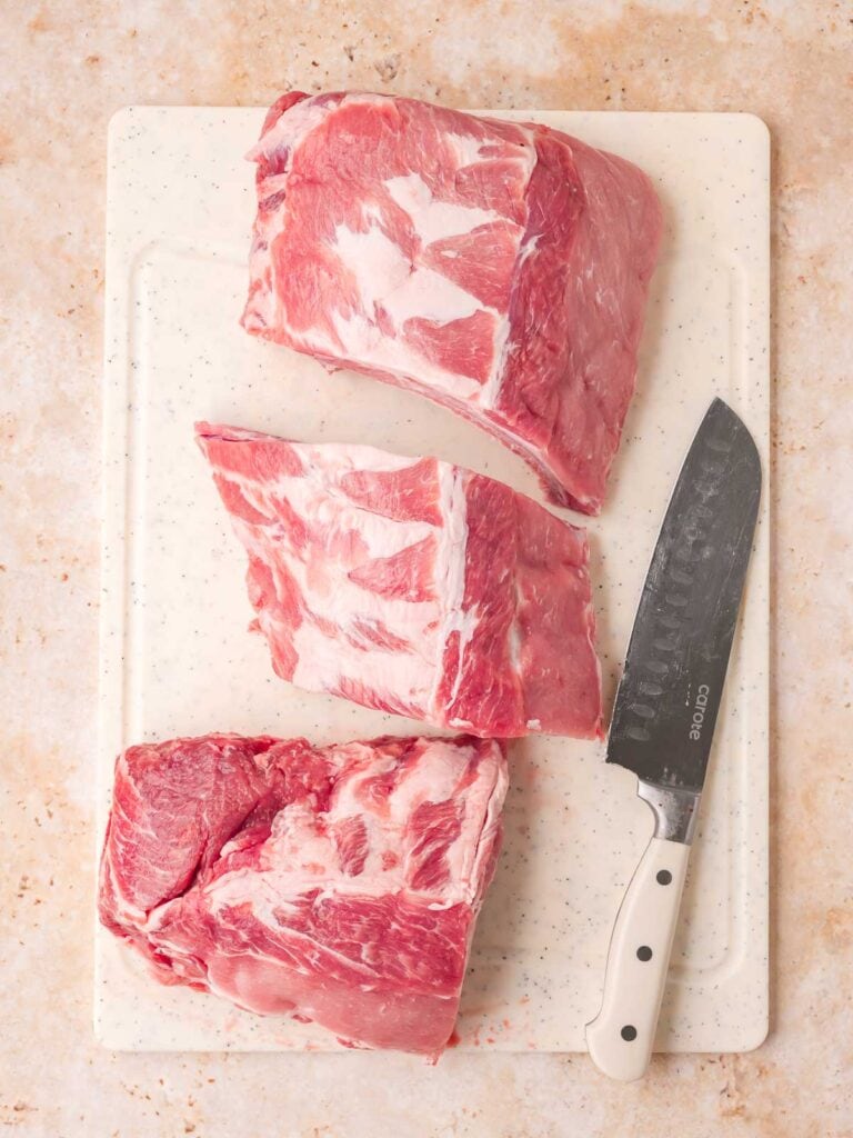 Raw pork spare ribs cut into three sections on a white cutting board with a knife — Fall-Off-the-Bone Oven BBQ Ribs by Tessie's Table.