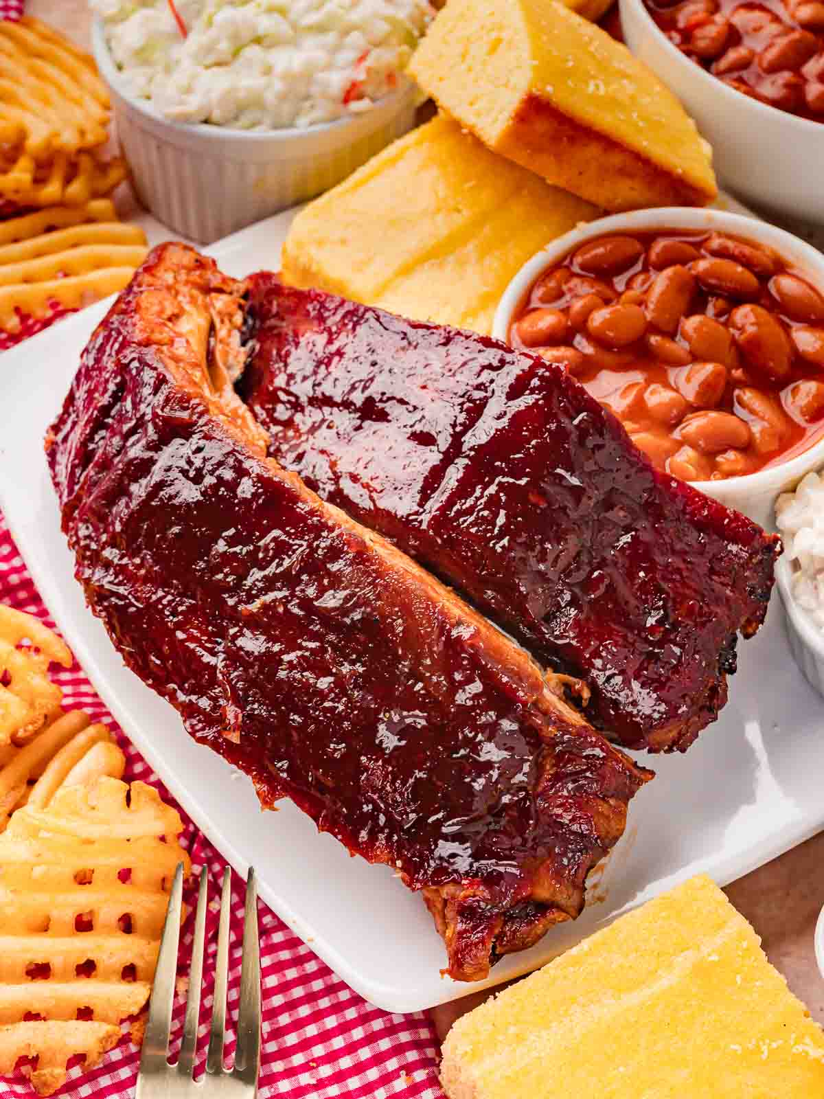 Fall-off-the-bone oven BBQ ribs on a white plate surrounded by waffle fries, cornbread, baked beans, and coleslaw — Fall-Off-the-Bone Oven BBQ Ribs by Tessie's Table.