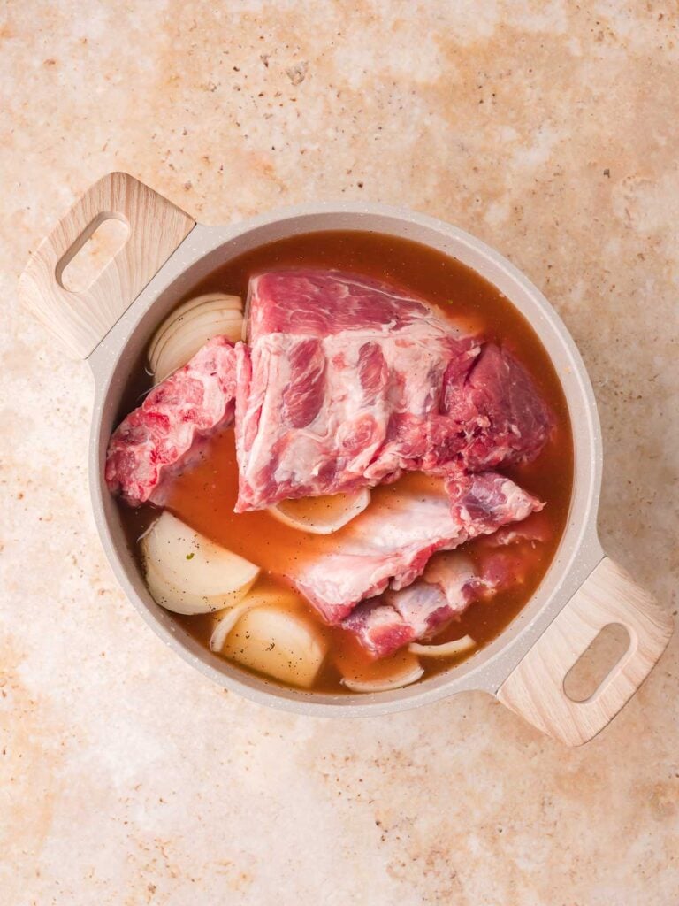 Raw pork spare rib sections in a large pot with chicken stock, quartered onion, and seasonings — Fall-Off-the-Bone Oven BBQ Ribs by Tessie's Table