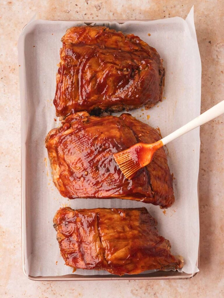 Cooked pork spare ribs on a parchment-lined baking tray being brushed with homemade BBQ sauce using a silicone pastry brush — Fall-Off-the-Bone Oven BBQ Ribs by Tessie's Table