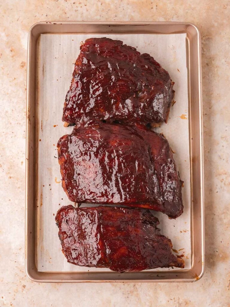 Three sections of oven baked BBQ ribs with sticky caramelized BBQ sauce on a parchment-lined baking tray — Fall-Off-the-Bone Oven BBQ Ribs by Tessie's Table