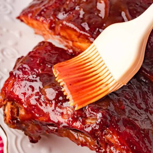 Close-up of a silicone pastry brush spreading homemade BBQ sauce over sticky glazed pork ribs on a white serving plate — Fall-Off-the-Bone Oven BBQ Ribs by Tessie's Table.