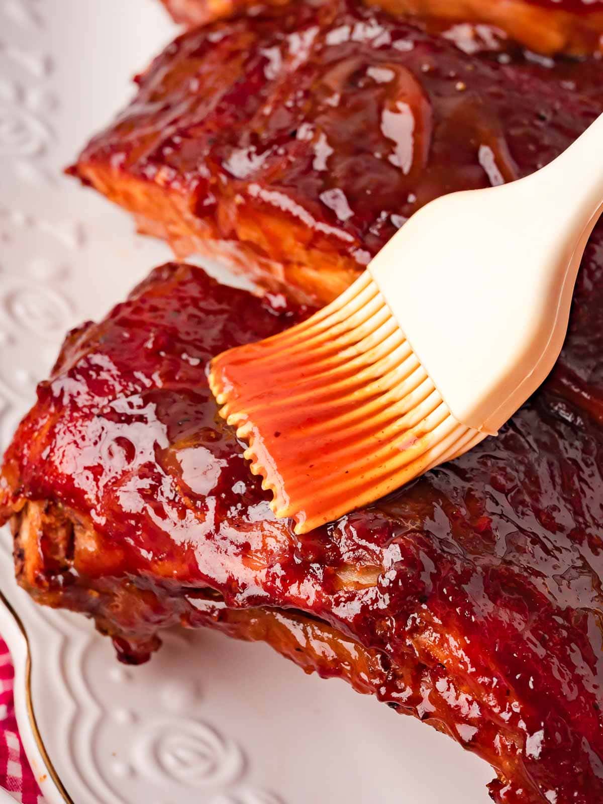 Close-up of a silicone pastry brush spreading homemade BBQ sauce over sticky glazed pork ribs on a white serving plate — Fall-Off-the-Bone Oven BBQ Ribs by Tessie's Table.