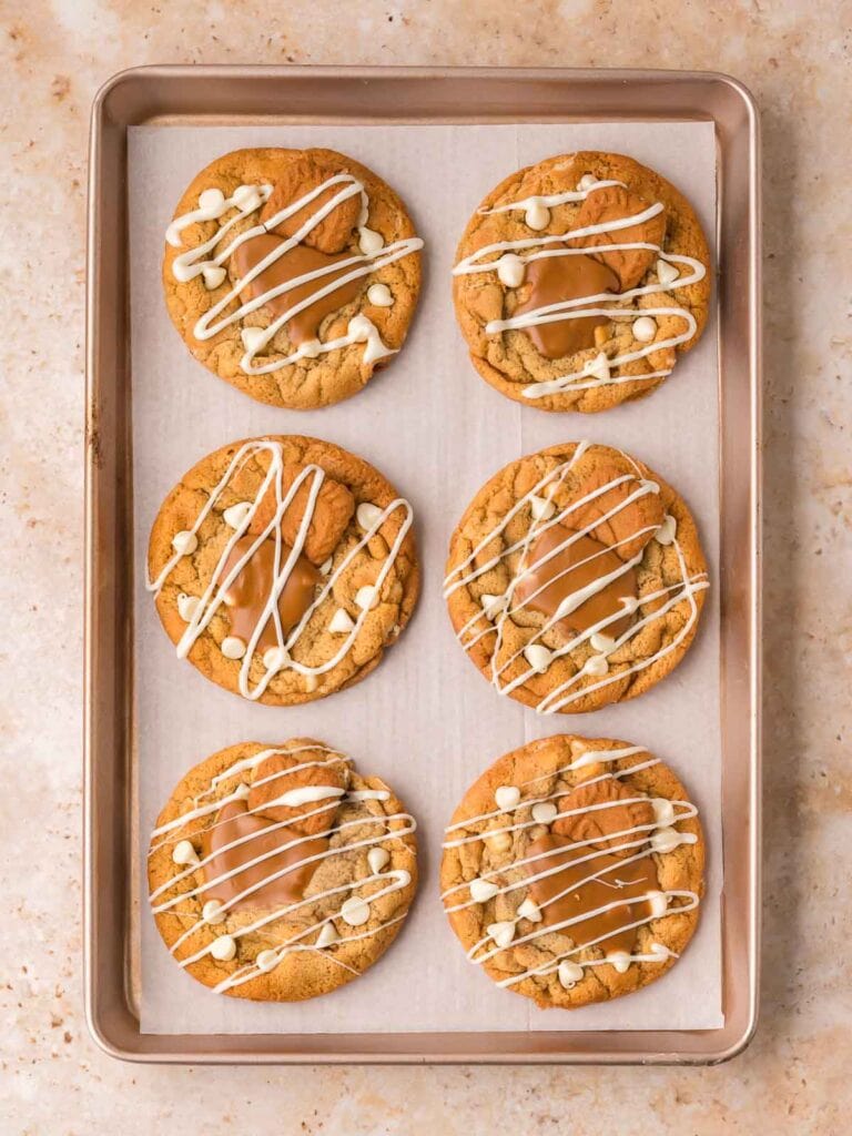 Six baked Biscoff stuffed cookies on a parchment-lined baking sheet with pooled Biscoff spread and white chocolate drizzle on top.