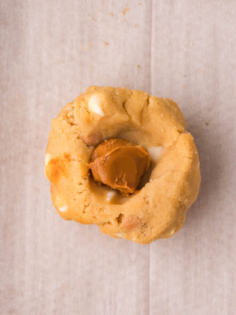 Flattened Biscoff stuffed cookie dough ball on parchment paper with a frozen Biscoff spread portion visible in the center before wrapping.