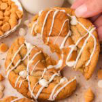 Hand breaking apart a Biscoff stuffed white chocolate cookie to reveal the gooey melted Biscoff center, with a glass of milk in the background.