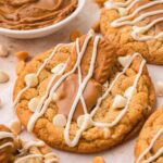 Close-up of a Biscoff stuffed white chocolate cookie topped with a Biscoff cookie and white chocolate drizzle, with a bowl of Biscoff spread in the background, by Tessie's Table.