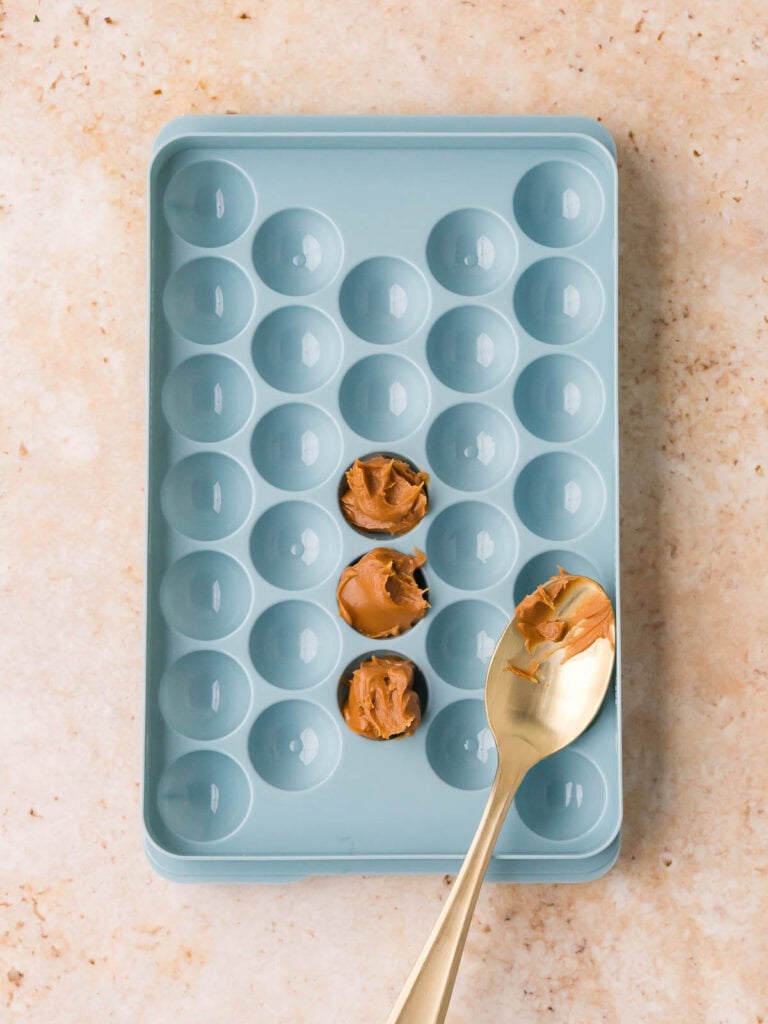 Biscoff spread being scooped into a blue silicone round mold with a gold spoon, three portions already filled, for Biscoff Stuffed White Chocolate Cookies by Tessie's Table.