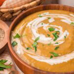 Wooden bowl of creamy homemade vegetable soup topped with cream and fresh parsley, with broccoli and carrots behind by Tessie's Table.