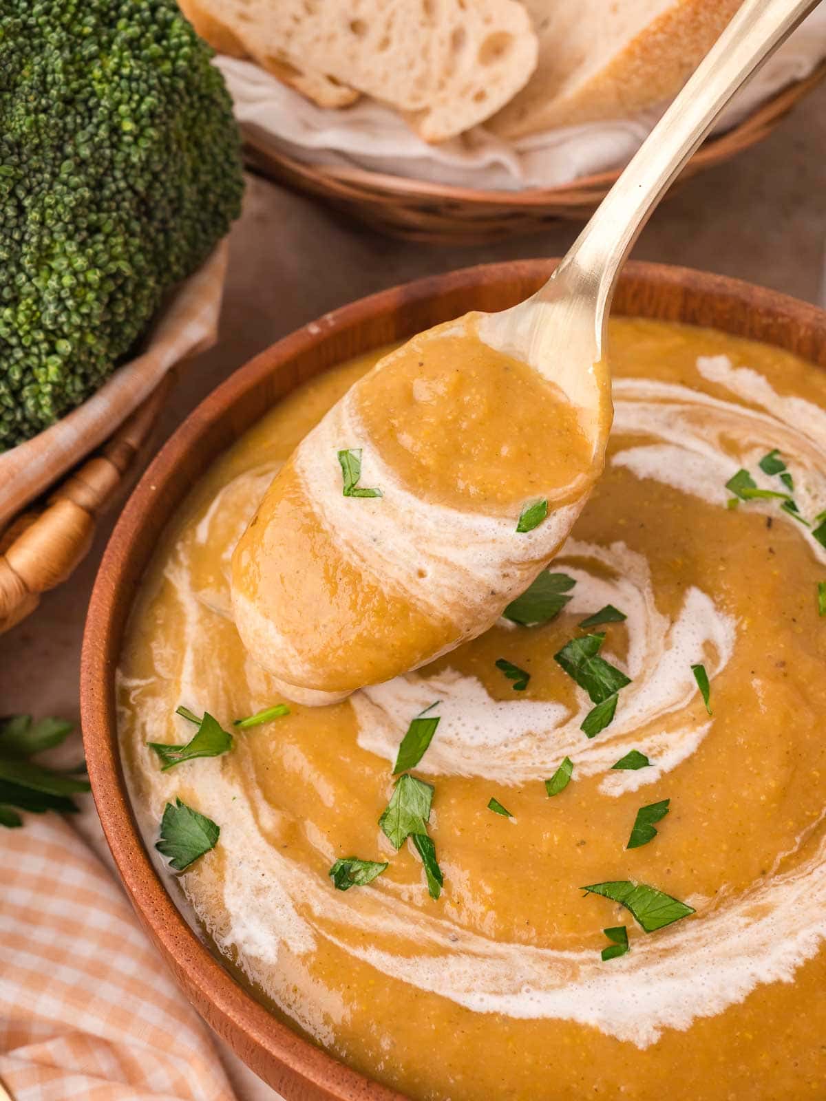 Gold spoon lifting a scoop of creamy homemade vegetable soup from a wooden bowl with bread and broccoli in the background by Tessie's Table.