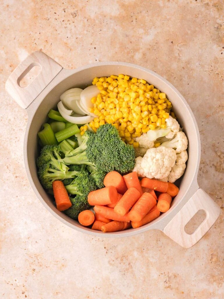 Raw broccoli, cauliflower, carrots, celery, onion, and sweet corn arranged in a soup pot for Creamy homemade vegetable soup by Tessie's Table.