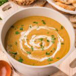 Pot of creamy homemade vegetable soup with a cream swirl, surrounded by baskets of bread, broccoli, and carrots by Tessie's Table.