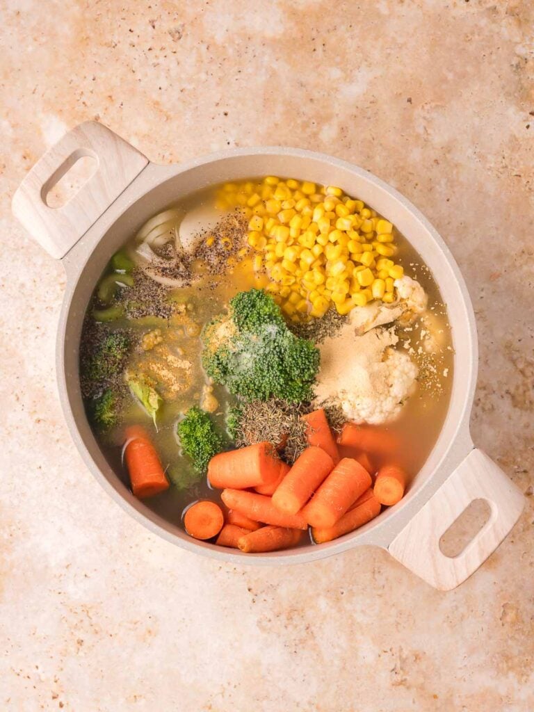 Chicken stock and seasonings added to broccoli, cauliflower, carrots, and corn in a pot for Creamy homemade vegetable soup by Tessie's Table.