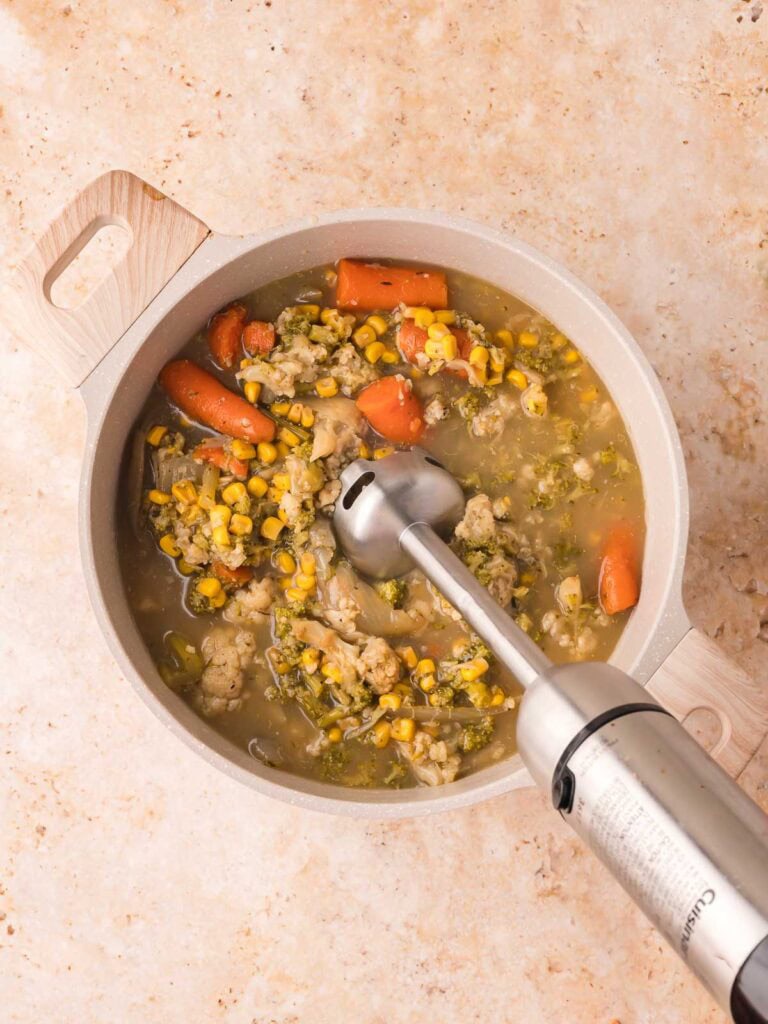 Hand blender pureeing softened vegetables into thick creamy homemade vegetable soup in a pot by Tessie's Table.