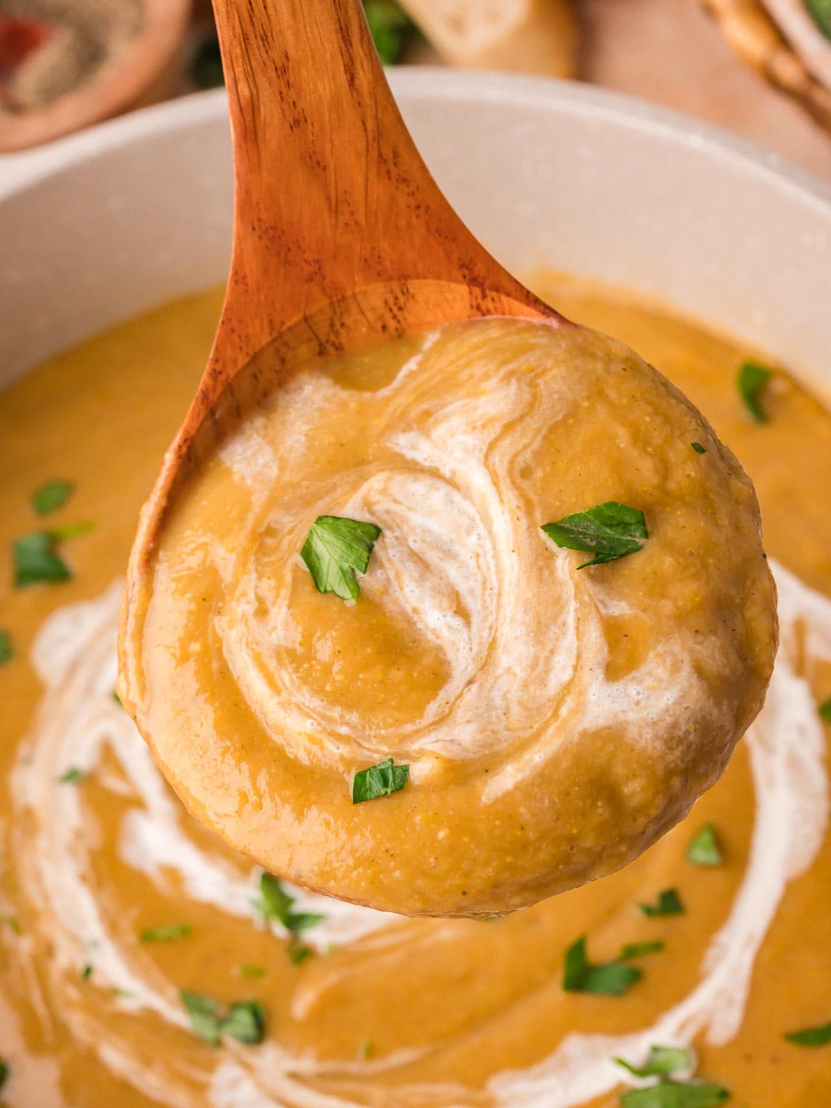 wooden spoon lifting a scoop of creamy homemade vegetable soup from a wooden bowl with bread and broccoli in the background by Tessie's Table.