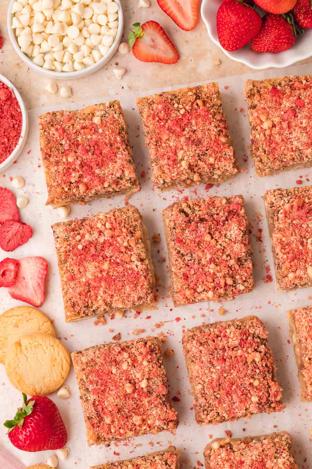 Strawberry crunch blondies arranged on parchment with white chocolate chips, freeze-dried strawberries, and fresh strawberries.
