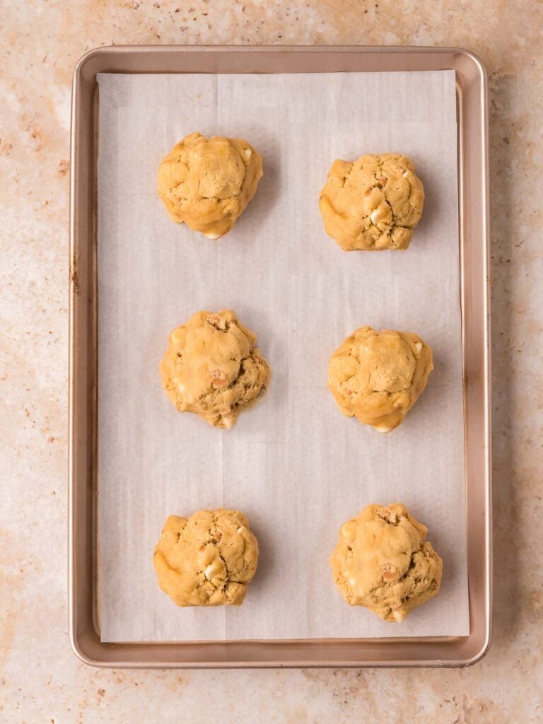 Six assembled Biscoff stuffed cookie dough balls on a parchment-lined baking sheet ready to go in the fridge to chill.