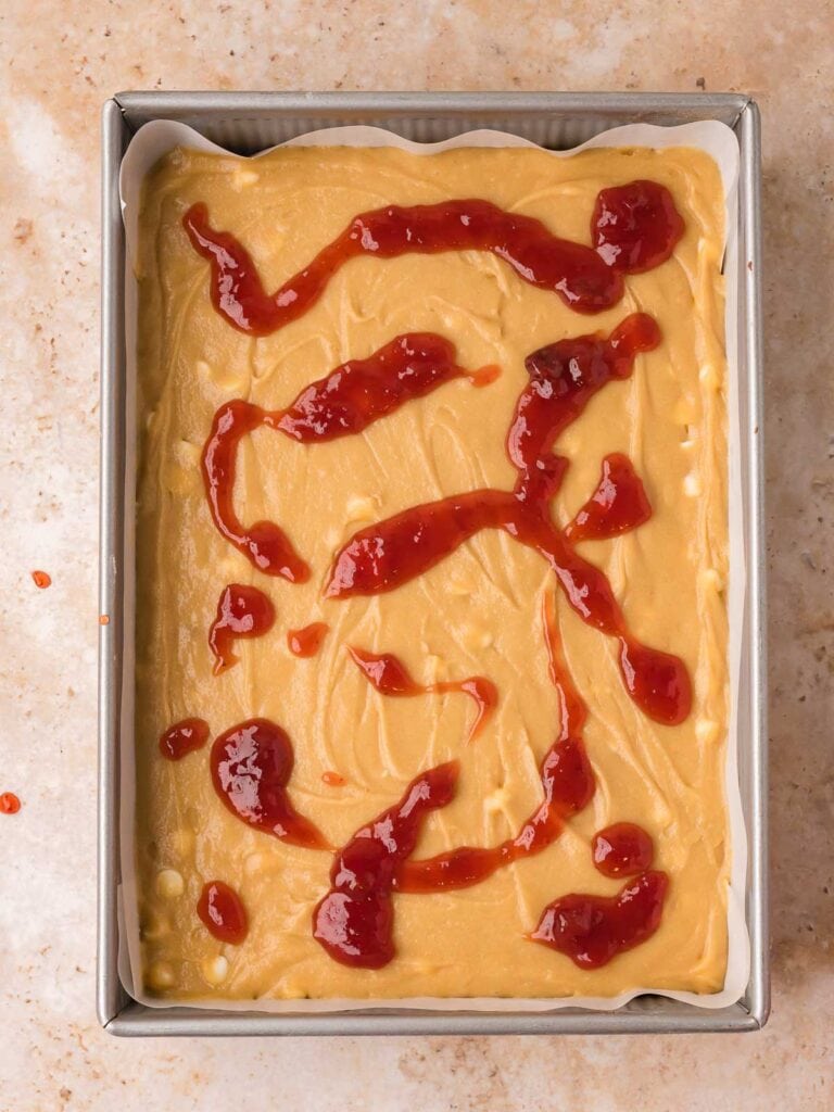 Strawberry jam dolloped over blondie batter spread in a parchment-lined pan.