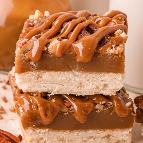 A stack of salted caramel pecan bars beside a jar of caramel and a glass of milk by Tessie's Table.