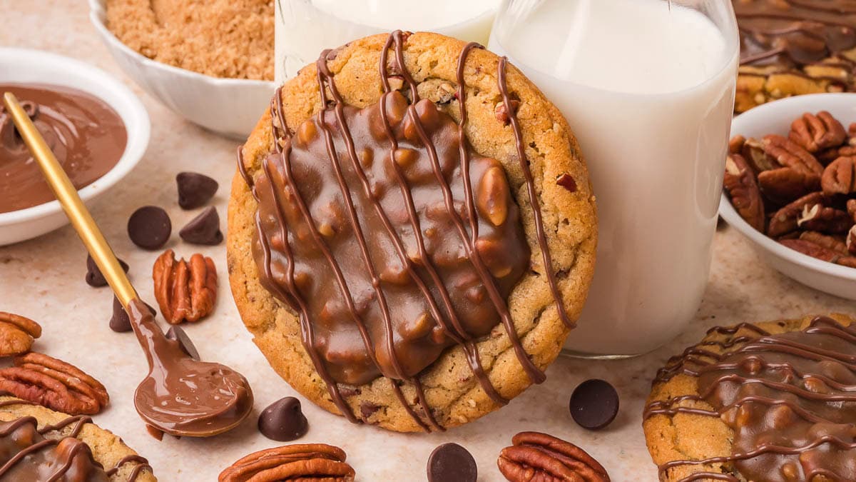 Pecan pie cookie topped with gooey pecan filling and chocolate drizzle next to a glass of milk by Tessie's Table.