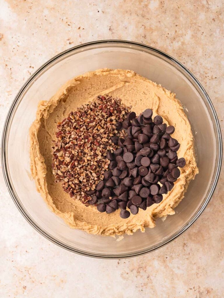 Chocolate chips and chopped pecans added to cookie dough in a glass bowl for pecan pie cookies by Tessie's Table.