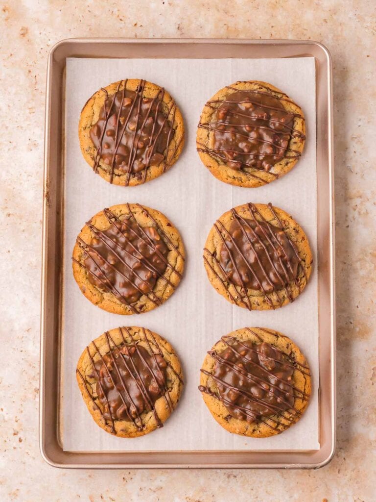 Six baked pecan pie cookies filled with pecan topping and chocolate drizzle on a baking sheet by Tessie's Table.