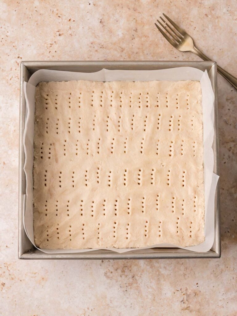 Shortbread dough pressed into a parchment lined square pan and pricked with a fork for salted caramel pecan bars by Tessie's Table.