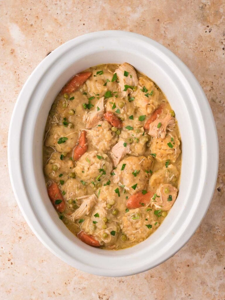Finished slow cooker chicken and dumplings with peas and carrots in a white crock by Tessie's Table.
